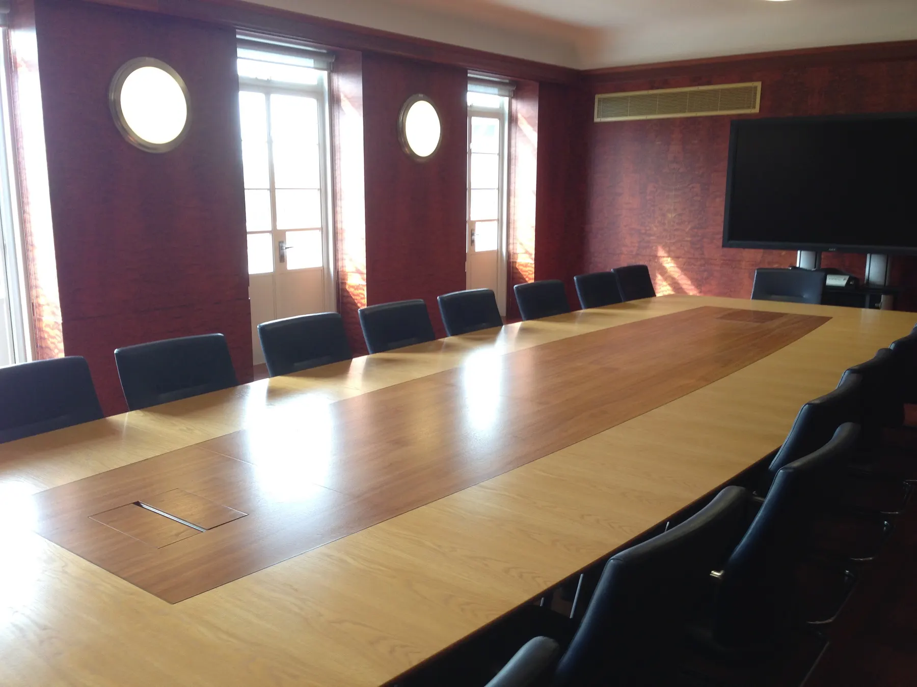 A long wooden meeting table lit by tall windows in a paneled room